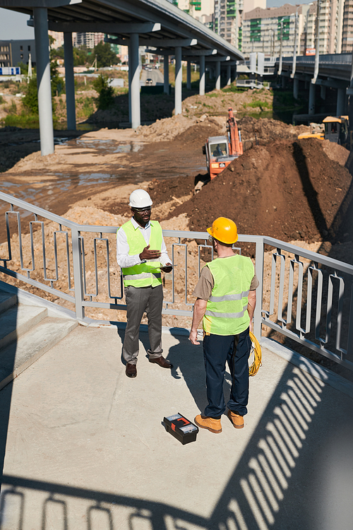 Backlit side view at two construction workers discussing project while standing at high rise building, copy space