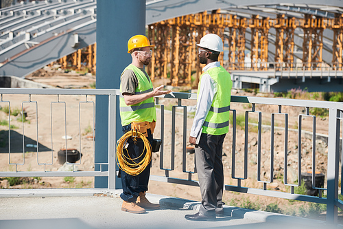 Engineers in hardhats and green vests discussing construction methods while viewing blueprint and standing on construction site with frames and cables