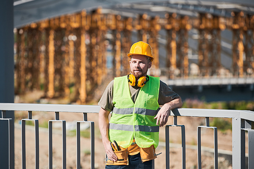 Waist up portrait of cheerful Middle-Eastern construction worker wearing harhat and holding metal bars smiling at camera while enjoying work on site