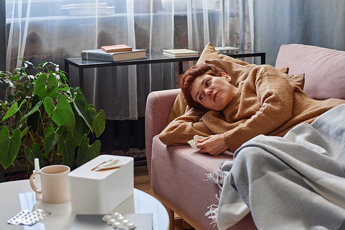 Horizontal shot of Caucasian woman having cold holding paper napkin lying on sofa at home