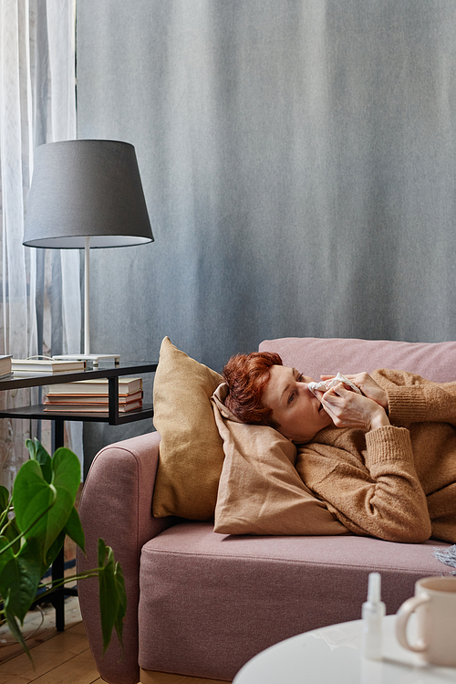 Vertical shot of Caucasian woman having influenza holding lying on sofa at home blowing her nose