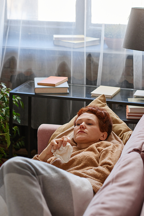 Vertical high angle shot of Caucasian woman suffering flu holding paper napkin relaxing on sofa at home