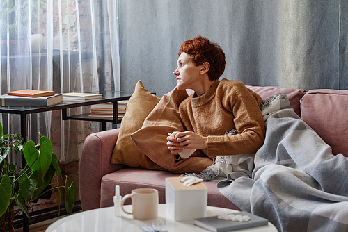 Horizontal shot of Caucasian woman suffering flu holding paper napkin relaxing on sofa at home looking away