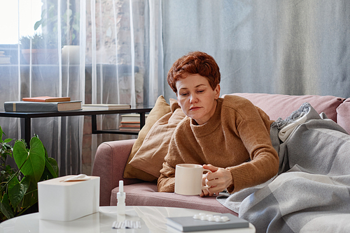 Horizontal shot of mature Caucasian woman with short hair feeling sick relaxing on sofa at home taking cup of water to take pills