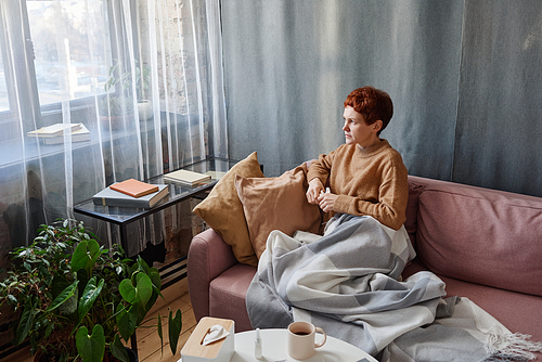 Horizontal high angle shot of mature Caucasian woman with short hair having coronavirus sitting on sofa at home looking away