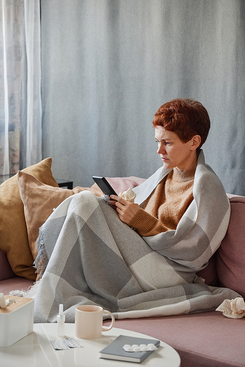 Vertical shot of mature Caucasian woman having coronavirus sitting on sofa wrapped in blanket surfing Internet on smartphone