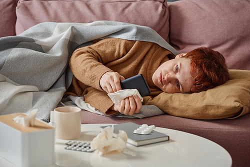 Horizontal medium shot of mature Caucasian woman having influenza relaxing on sofa surfing Internet on smartphone