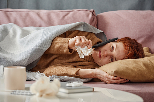 Horizontal portrait of mature Caucasian woman having influenza lying on sofa talking on phone with someone