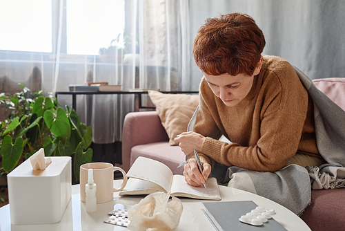 Horizontal shot of mature businesswoman having flu staying at home making notes in notebook