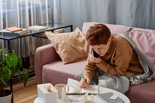 Horizontal shot of mature woman suffering flu staying at home talking to doctor on phone making notes in notebook