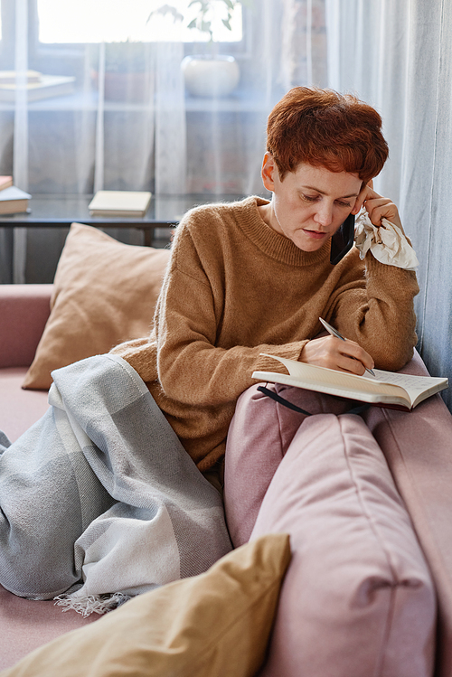 Vertical shot of modern mature woman having influenza staying at home talking to doctor on phone writing something in notebook