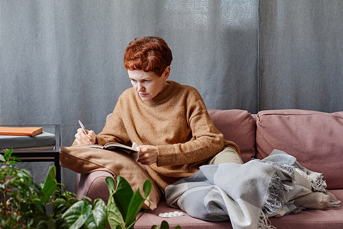 Horizontal shot of mature Caucasian woman having cold staying at home working on something making notes in notebook