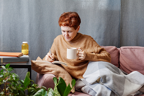 Horizontal shot of mature Caucasian woman having influenza staying at home working on something making notes in notebook and drinking tea