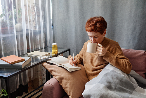 High angle shot of modern mature Caucasian woman having influenza staying at home sitting on sofa writing notes in notebook and drinking tea