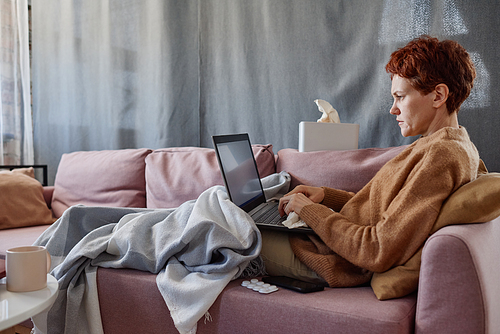 Horizontal shot of mature Caucasian woman suffering cold staying at home texting something on laptop