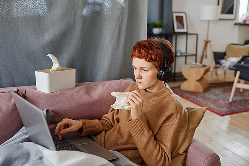 Horizontal shot of mature Caucasian woman having influenza staying at home sitting on sofa watching something on laptop