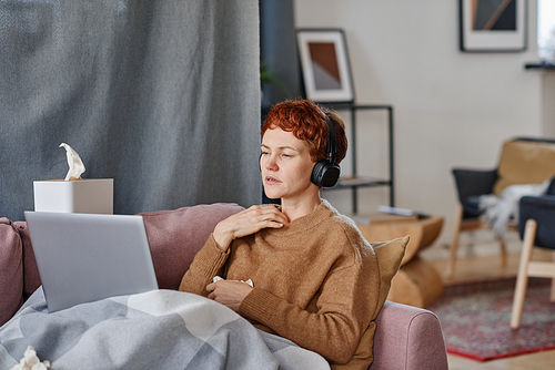 Horizontal shot of mature Caucasian woman having influenza staying at home sitting on sofa having doctors appointment online