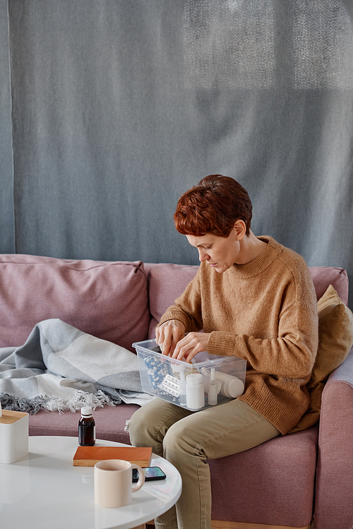 Vertical medium long shot of modern mature Caucasian woman sitting on sofa with first aid kit on her lap