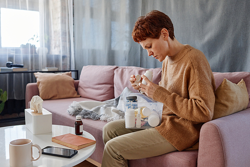 Horizontal medium shot of modern mature Caucasian woman sitting on sofa with first aid kit on her lap looking for pills to treat flu symptoms