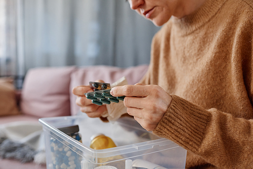 Horizontal medium close-up shot of mature Caucasian woman sitting on sofa with first aid kit on her lap choosing pills to treat headache