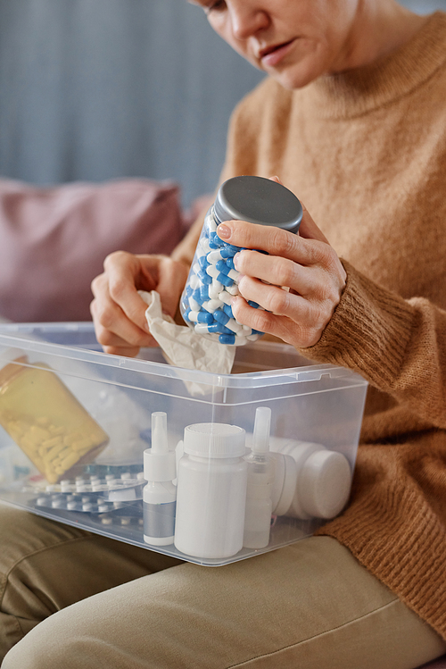 Vertical medium section shot of mature woman suffering headache sitting on sofa with first aid kit on her lap choosing pills to take