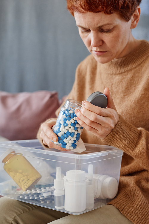 Vertical medium shot of mature woman suffering flu symptoms sitting on sofa with first aid kit on her lap opening jar with meds in it