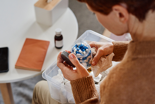 High angle medium close-up shot of unrecognizable woman suffering coronavirus sitting on sofa with first aid kit on her lap opening jar with pills in it