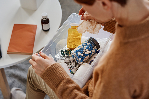 High angle medium close-up shot of unrecognizable woman suffering fever sitting on sofa with first aid kit on her lap looking for pills