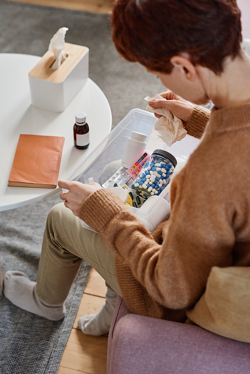 Vertical high angle shot of woman suffering influenza sitting on sofa with first aid kit on her lap looking for pills to take