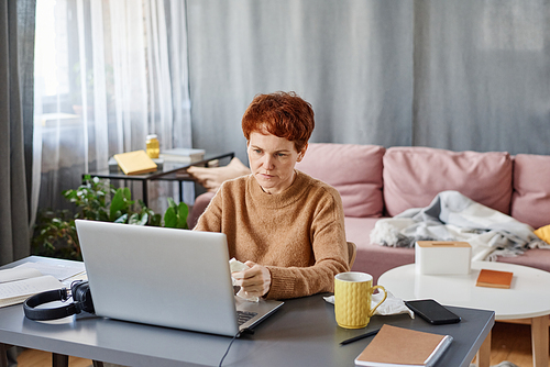 Horizontal shot of mature Caucasian woman suffering cold staying at home sitting at desk working on laptop