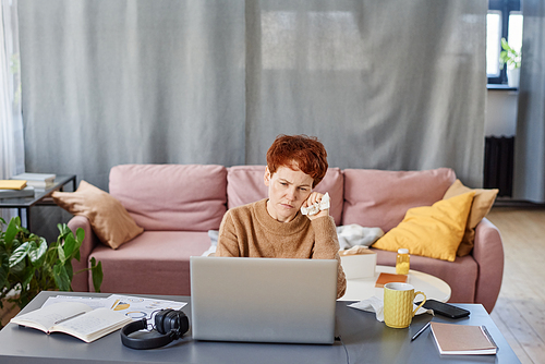 Horizontal shot of mature Caucasian woman suffering influenza staying at home sitting at desk working on laptop