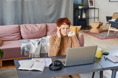 Horizontal shot of mature Caucasian woman suffering fever with headache staying at home sitting at desk working on laptop