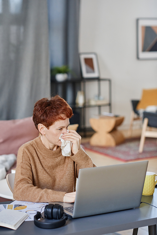 Vertical shot of mature Caucasian woman suffering flu staying at home sitting at desk working on laptop and blowing her nose