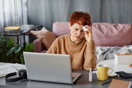 Horizontal shot of mature Caucasian woman sitting at desk at home in front of laptop measuring body temperature using thermometer