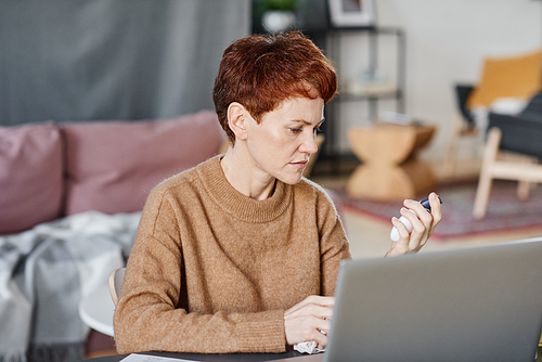 Horizontal medium close-up shot of mature Caucasian woman sitting at desk at home in front of laptop checking body temperature using thermometer