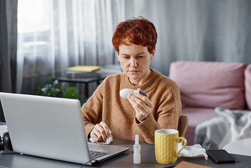 Portrait shot of mature Caucasian woman having influenza sitting at desk at home in front of laptop checking body temperature using thermometer