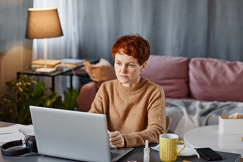 Portrait of mature Caucasian woman having flu staying at home sitting at desk watching content in Internet using laptop