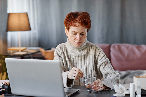 Mature woman having influenza wearing cozy sweater sitting at desk at home in front of laptop putting carbon tablet into glass of water