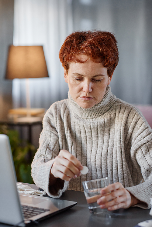 Vertical portrait of mature woman having influenza wearing sweater sitting at desk at home putting effervescent tablet into glass of water