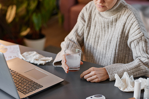 High angle shot of unrecognizable woman having flu wearing sweater sitting at desk at home holding glass with mixture of water and effervescent tablet