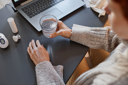 High angle shot of woman having cold wearing sweater sitting at desk at home holding glass with mixture of water