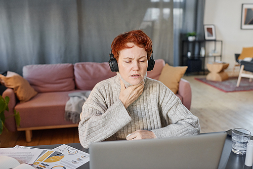 Horizontal shot of mature woman suffering flu with sore throat wearing headphones sitting at desk at home touching neck