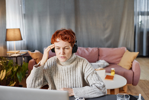 Mature woman sitting at desk in living room at home telling physician about headache during online consultation