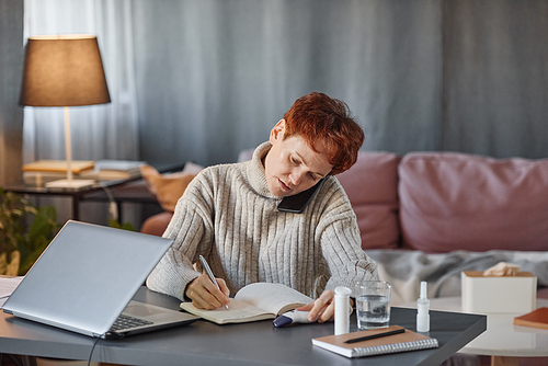 Mature woman having influenza sitting at desk in living room at home talking on phone with colleague and making notes