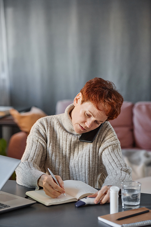 Vertical shot of mature woman having influenza sitting at desk in living room at home talking on phone making appointment with doctor