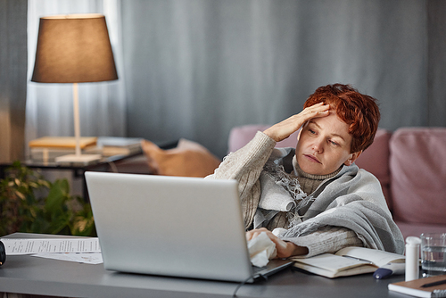 Portrait of mature Caucasian woman having flu with severe headache wearing warm outfit sitting at desk in living room at home trying to work