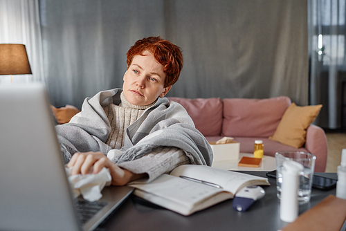 Portrait of mature Caucasian woman having coronavirus wearing warm outfit sitting at desk in living room at home watching something on laptop