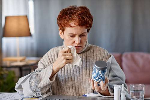 Horizontal portrait shot of mature Caucasian woman sitting at desk in front of laptop having online appointment looking at jar full of vitamin pills