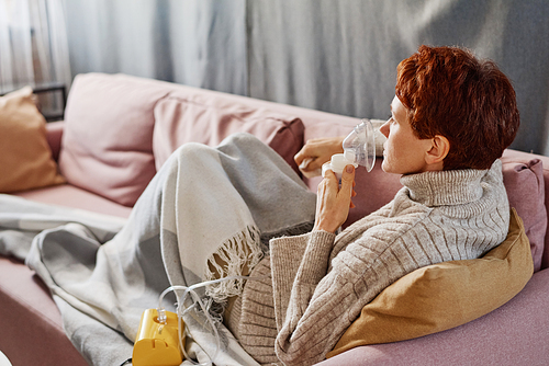 Horizontal medium shot of modern mature Caucasian woman with short red hair having influenza relaxing at home turning on nebulizer