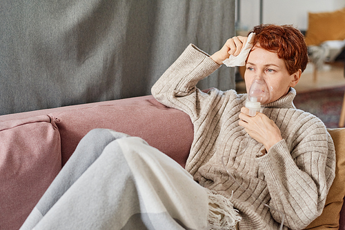 Horizontal medium shot of mature Caucasian woman with short red hair having influenza suffering fever and cough relaxing at home using nebulizer